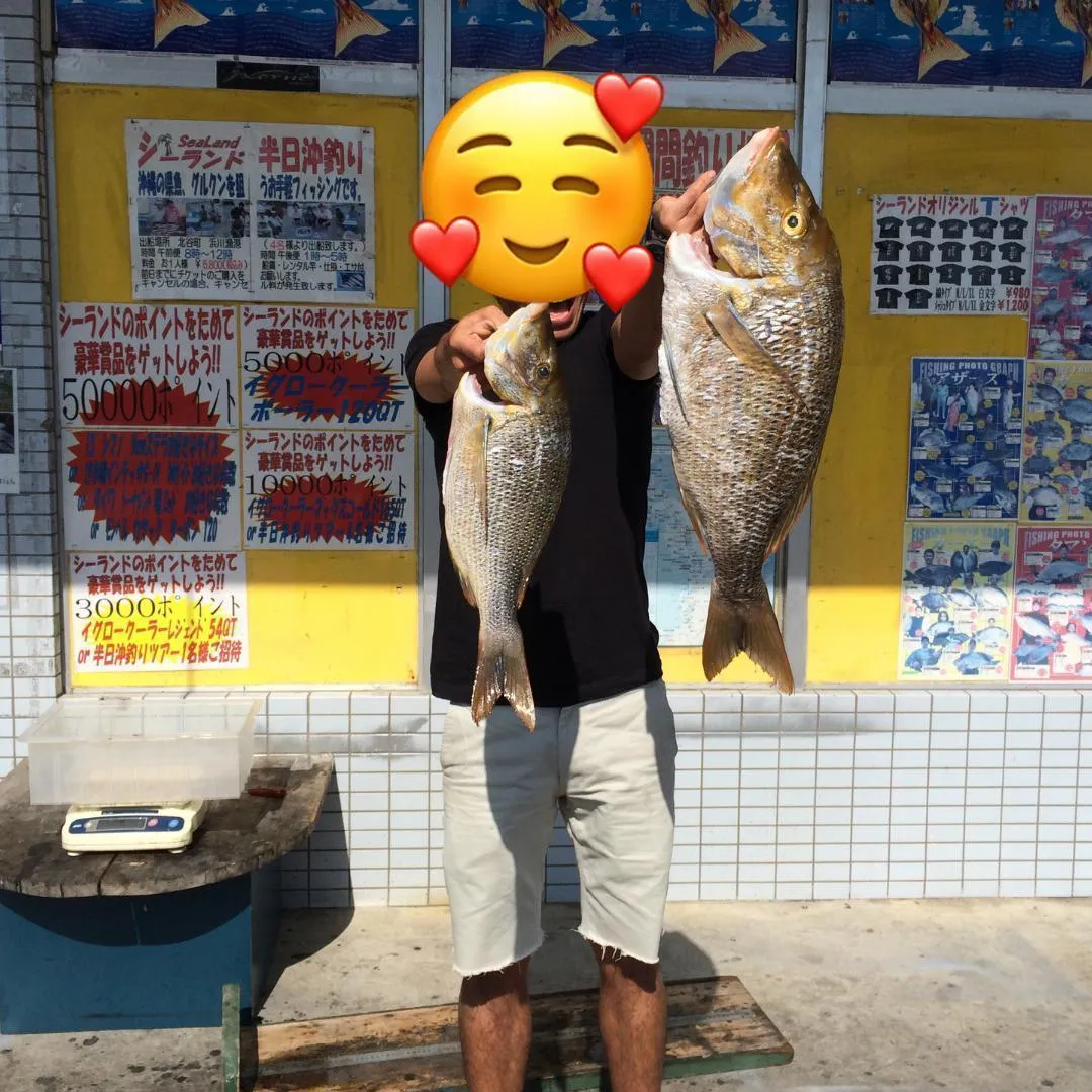 黄色い掲示板の前で魚を2匹持つ人（顔は絵文字で隠している）